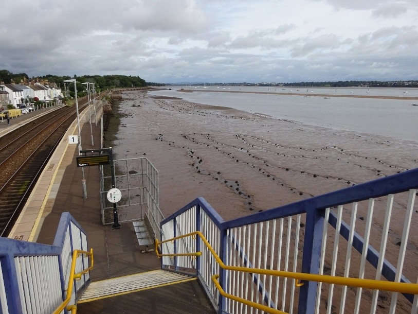 A photo taken from the bridge over the railway tracks at Starcross train station in Devon. The platform is to the left, immediately next to it is an expanse of exposed mud, with the Exe following beyond it. It's a grey day, boats can be seen on the river.